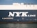 The side view of a large cargo ship with the words 'NYK LINE' prominently displayed in large white letters on a dark blue background. The ocean is visible at the bottom edge of the image, reflecting the ship's hull.