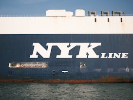 The side view of a large cargo ship with the words 'NYK LINE' prominently displayed in large white letters on a dark blue background. The ocean is visible at the bottom edge of the image, reflecting the ship's hull.