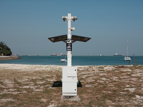 A beach landscape with a metal structure resembling a solar panel station in the center foreground. The background features a calm blue sea with several boats floating, and a clear blue sky. The sandy beach has patches of grass and a tree-lined shore on the left side.