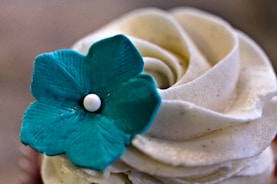 A beautifully swirled white frosting with a single blue decorative flower placed on top, creating a visually appealing contrast. The frosting has a smooth texture and the flower features intricate petal details with a small white center.