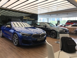 A modern car showroom featuring several luxury vehicles. A vibrant blue car is prominently displayed in the foreground, while other cars in black and silver are positioned behind it. The showroom has a sleek, minimalist design with a large window allowing natural light to illuminate the space. There are chairs and a backpack visible in the foreground.