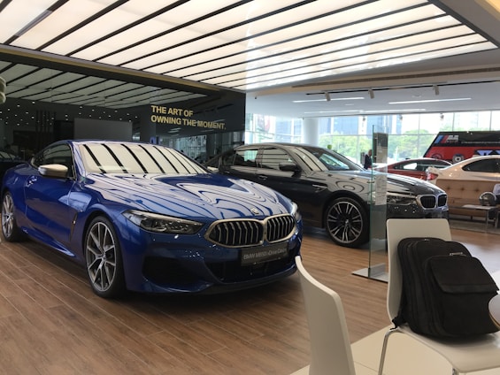 A modern car showroom featuring several luxury vehicles. A vibrant blue car is prominently displayed in the foreground, while other cars in black and silver are positioned behind it. The showroom has a sleek, minimalist design with a large window allowing natural light to illuminate the space. There are chairs and a backpack visible in the foreground.