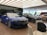 A modern car showroom featuring several luxury vehicles. A vibrant blue car is prominently displayed in the foreground, while other cars in black and silver are positioned behind it. The showroom has a sleek, minimalist design with a large window allowing natural light to illuminate the space. There are chairs and a backpack visible in the foreground.