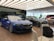 A modern car showroom featuring several luxury vehicles. A vibrant blue car is prominently displayed in the foreground, while other cars in black and silver are positioned behind it. The showroom has a sleek, minimalist design with a large window allowing natural light to illuminate the space. There are chairs and a backpack visible in the foreground.