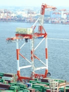 A large industrial crane stands prominently over a bustling port filled with shipping containers. The crane features a striking white and orange color scheme, and the containers below are marked with 'Evergreen'. In the background, multiple cranes and industrial structures are visible along the waterfront.