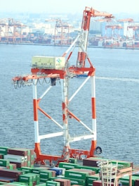 A large industrial crane stands prominently over a bustling port filled with shipping containers. The crane features a striking white and orange color scheme, and the containers below are marked with 'Evergreen'. In the background, multiple cranes and industrial structures are visible along the waterfront.