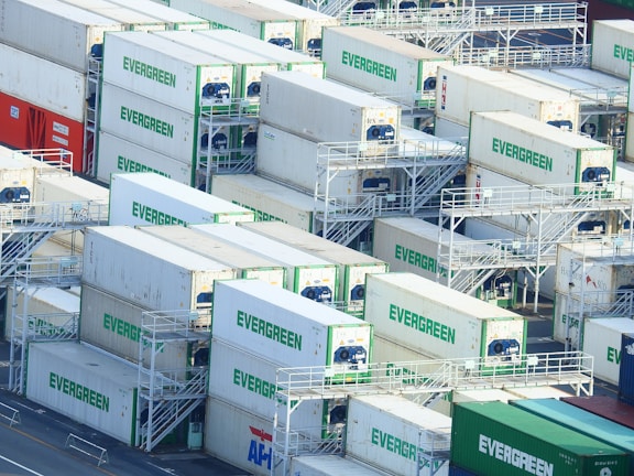 A densely packed arrangement of shipping containers, predominantly white with the word 'EVERGREEN' in green, stacked in an orderly fashion at a shipping port. The containers are on racks and appear to be refrigerated, with visible cooling units. A few containers in red, blue, and green contrast with the majority.