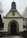 A small chapel with a grey, shingled steeple and beige walls is situated in a cemetery. The entrance has an arched wooden door framed by a carved inscription in German. Sparse snow covers the ground, and bare trees are visible in the background.