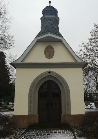 A small chapel with a grey, shingled steeple and beige walls is situated in a cemetery. The entrance has an arched wooden door framed by a carved inscription in German. Sparse snow covers the ground, and bare trees are visible in the background.