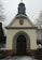 A small chapel with a grey, shingled steeple and beige walls is situated in a cemetery. The entrance has an arched wooden door framed by a carved inscription in German. Sparse snow covers the ground, and bare trees are visible in the background.
