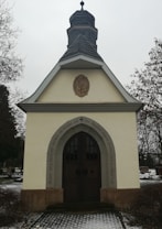 A small chapel with a grey, shingled steeple and beige walls is situated in a cemetery. The entrance has an arched wooden door framed by a carved inscription in German. Sparse snow covers the ground, and bare trees are visible in the background.