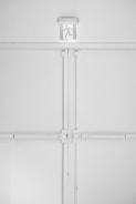 A minimalist emergency exit sign is mounted above a large, industrial-style set of double doors. The sign features a pictogram of a person running, suggesting direction for evacuation. The entire scene is set against a predominantly white background with some subtle shadows and textures created by the doors and frames.