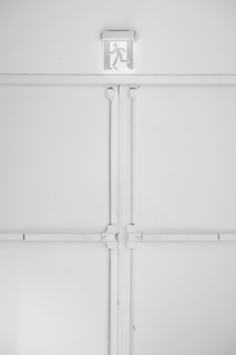 A minimalist emergency exit sign is mounted above a large, industrial-style set of double doors. The sign features a pictogram of a person running, suggesting direction for evacuation. The entire scene is set against a predominantly white background with some subtle shadows and textures created by the doors and frames.
