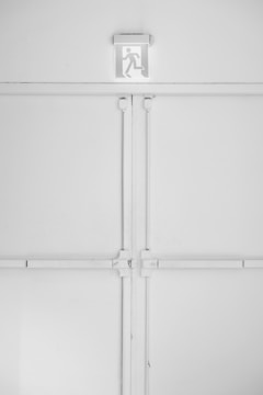 A minimalist emergency exit sign is mounted above a large, industrial-style set of double doors. The sign features a pictogram of a person running, suggesting direction for evacuation. The entire scene is set against a predominantly white background with some subtle shadows and textures created by the doors and frames.