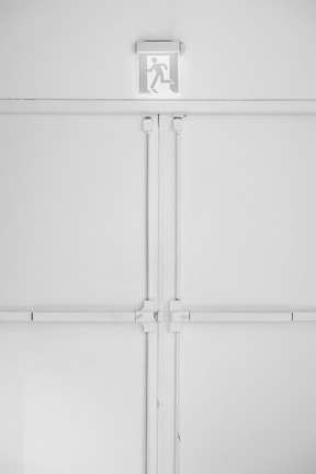 A minimalist emergency exit sign is mounted above a large, industrial-style set of double doors. The sign features a pictogram of a person running, suggesting direction for evacuation. The entire scene is set against a predominantly white background with some subtle shadows and textures created by the doors and frames.