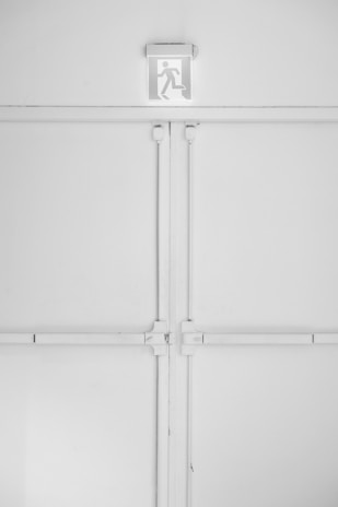 A minimalist emergency exit sign is mounted above a large, industrial-style set of double doors. The sign features a pictogram of a person running, suggesting direction for evacuation. The entire scene is set against a predominantly white background with some subtle shadows and textures created by the doors and frames.