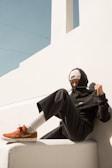A person dressed in a black hoodie and pants is sitting casually on a white structure. They are wearing a white knitted cap and adjusting the hoodie string. The scene is set against a backdrop of a minimalist architectural wall with a bright blue sky overhead. The style is urban and modern.