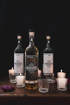 Three elegant glass bottles prominently feature intricate labels with the words 'Dos Conejo.' They are arranged on a wooden surface surrounded by several lit votive candles in glass holders. The lighting is dim, creating a warm atmosphere.