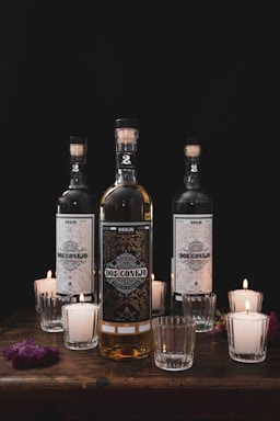 Three elegant glass bottles prominently feature intricate labels with the words 'Dos Conejo.' They are arranged on a wooden surface surrounded by several lit votive candles in glass holders. The lighting is dim, creating a warm atmosphere.
