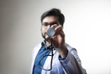 A male doctor wearing glasses and a white coat holds a stethoscope towards the camera. The background is plain and light-colored, which makes the doctor the central focus of the image. The man appears to be looking directly at the camera through the stethoscope.