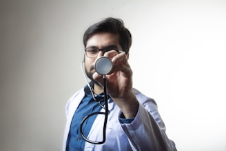A male doctor wearing glasses and a white coat holds a stethoscope towards the camera. The background is plain and light-colored, which makes the doctor the central focus of the image. The man appears to be looking directly at the camera through the stethoscope.