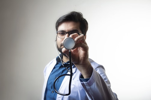 A male doctor wearing glasses and a white coat holds a stethoscope towards the camera. The background is plain and light-colored, which makes the doctor the central focus of the image. The man appears to be looking directly at the camera through the stethoscope.