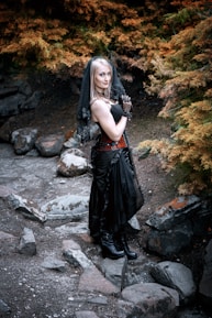 A woman dressed in a gothic-style outfit stands outdoors, surrounded by autumnal foliage. She wears a black dress with lace gloves, a veil, and black boots, standing on a rocky path amidst burnt orange and green trees.