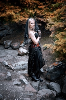 A woman dressed in a gothic-style outfit stands outdoors, surrounded by autumnal foliage. She wears a black dress with lace gloves, a veil, and black boots, standing on a rocky path amidst burnt orange and green trees.