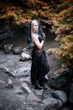 A woman dressed in a gothic-style outfit stands outdoors, surrounded by autumnal foliage. She wears a black dress with lace gloves, a veil, and black boots, standing on a rocky path amidst burnt orange and green trees.