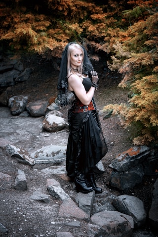 A woman dressed in a gothic-style outfit stands outdoors, surrounded by autumnal foliage. She wears a black dress with lace gloves, a veil, and black boots, standing on a rocky path amidst burnt orange and green trees.