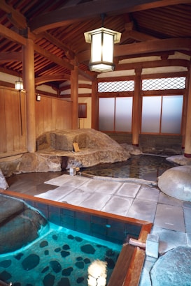A traditional Japanese onsen bathhouse setting with wooden architecture and rock features. The interior includes a pool of clear water with visible stones at the bottom. The area is illuminated by hanging lanterns, reflecting a serene and calm ambiance. Wooden beams and frosted glass windows add to the authentic aesthetic.