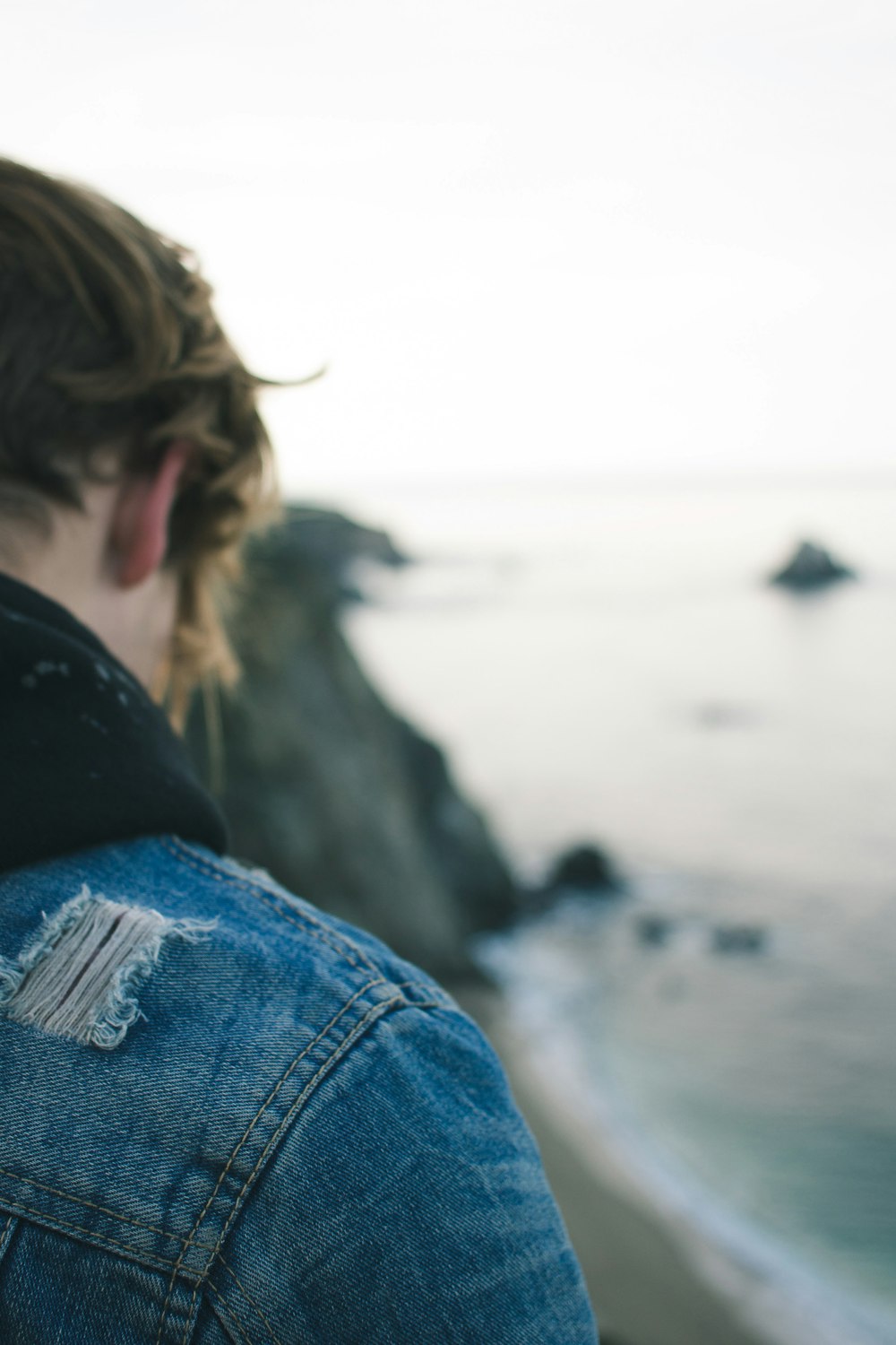 Frau in blauer Jeansjacke und blauer Jeans sitzt auf Felsen in der Nähe von Wasser