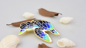 A colorful enamel pin in the shape of a sea turtle featuring shades of blue, green, and gold. The pin is placed on a light, neutral background along with several white seashells and a few brown dried leaves scattered around.