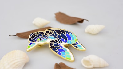 A colorful enamel pin in the shape of a sea turtle featuring shades of blue, green, and gold. The pin is placed on a light, neutral background along with several white seashells and a few brown dried leaves scattered around.