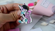 A hand holding a colorful pin depicting a cartoon animal character with black hair, pink and white spotted fur, and bright blue accents on its paws and eyes. The background includes a blurred pink paper wrapped bouquet and white surface with some scattered items.
