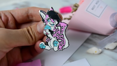 A hand holding a colorful pin depicting a cartoon animal character with black hair, pink and white spotted fur, and bright blue accents on its paws and eyes. The background includes a blurred pink paper wrapped bouquet and white surface with some scattered items.