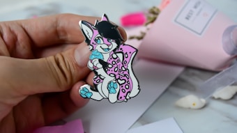 A hand holding a colorful pin depicting a cartoon animal character with black hair, pink and white spotted fur, and bright blue accents on its paws and eyes. The background includes a blurred pink paper wrapped bouquet and white surface with some scattered items.