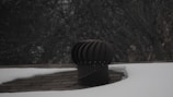 A rooftop vent is surrounded by snow with a dark, leafless tree in the background. The vent is cylindrical with a ridged top, appearing metallic in nature. The shingles on the roof are partially visible where the snow has not covered them.