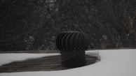 A rooftop vent is surrounded by snow with a dark, leafless tree in the background. The vent is cylindrical with a ridged top, appearing metallic in nature. The shingles on the roof are partially visible where the snow has not covered them.