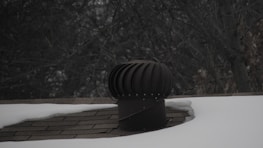 A rooftop vent is surrounded by snow with a dark, leafless tree in the background. The vent is cylindrical with a ridged top, appearing metallic in nature. The shingles on the roof are partially visible where the snow has not covered them.