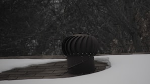 A rooftop vent is surrounded by snow with a dark, leafless tree in the background. The vent is cylindrical with a ridged top, appearing metallic in nature. The shingles on the roof are partially visible where the snow has not covered them.