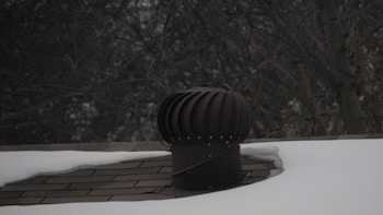 A rooftop vent is surrounded by snow with a dark, leafless tree in the background. The vent is cylindrical with a ridged top, appearing metallic in nature. The shingles on the roof are partially visible where the snow has not covered them.