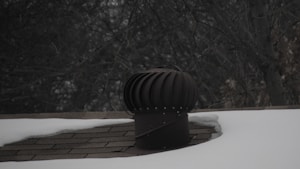 A rooftop vent is surrounded by snow with a dark, leafless tree in the background. The vent is cylindrical with a ridged top, appearing metallic in nature. The shingles on the roof are partially visible where the snow has not covered them.