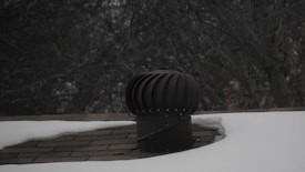 A rooftop vent is surrounded by snow with a dark, leafless tree in the background. The vent is cylindrical with a ridged top, appearing metallic in nature. The shingles on the roof are partially visible where the snow has not covered them.