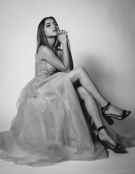 A woman in an elegant gown sits gracefully with her legs crossed and her hands clasped together under her chin. The gown is embellished with intricate beadwork and flows delicately around her. Her expression is contemplative, exuding poise and confidence.