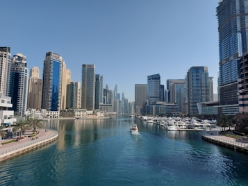 A cityscape featuring a series of modern skyscrapers lining both sides of a wide water channel. Several yachts are moored along the docks, and a boat is traveling down the center of the waterway. The sky is clear, and there are palm trees visible along the promenade.