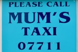 A sign with bold blue text on a lighter blue background that reads 'PLEASE CALL MUM'S TAXI 07711'. The text is aligned centrally and uses capital letters.