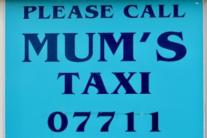 A sign with bold blue text on a lighter blue background that reads 'PLEASE CALL MUM'S TAXI 07711'. The text is aligned centrally and uses capital letters.