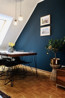 A modern dining room features a wooden table with black wire chairs, complemented by two vases filled with yellow flowers. The room has a dark blue accent wall with two framed architectural prints. A small citrus tree in a stylish woven basket adds a natural touch to the decor. Three spherical pendant lights hang above the table, creating a cozy ambiance.