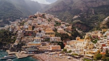 Colorful hillside village overlooking the coastline, with numerous tightly packed buildings rising up against a backdrop of steep, green mountains. The foreground features a sandy beach with beachgoers and a clear blue sea.